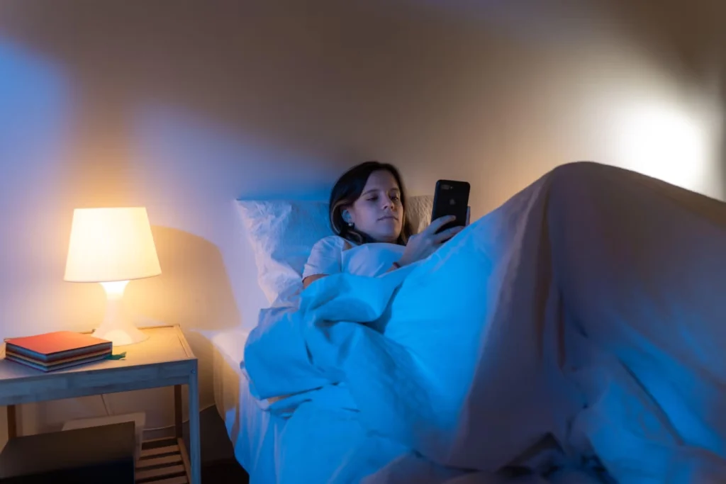 Person sitting mentally exhausted on a bed at night while doom scrolling on a phone
