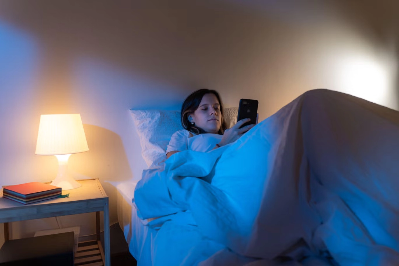 Person sitting mentally exhausted on a bed at night while doom scrolling on a phone